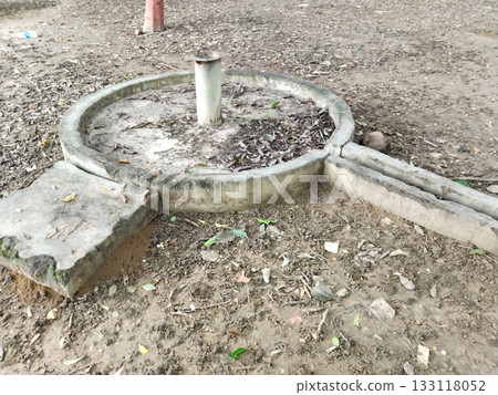 Old concrete water handpump base in park, circular structure with pipe and drainage, 133118052