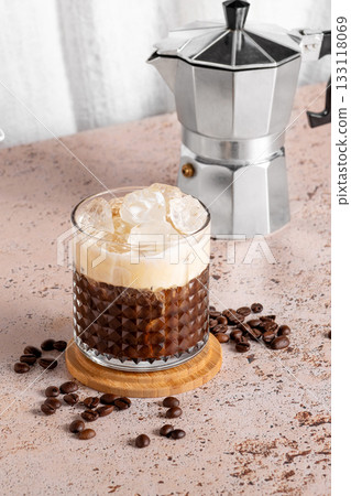 Iced creamy coffee into a glass with moka pot on beige kitchen table 133118069
