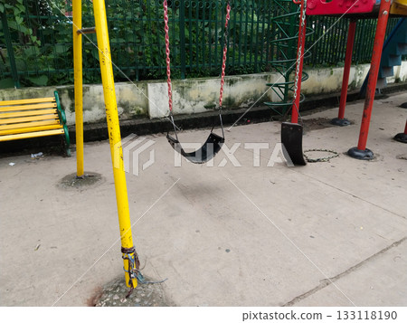 Black rubber swings hanging from chains in public park playground, simple outdoor play equipment for children, placed on concrete ground with yellow and red frames, offering fun activity and classic Black rubber swings hanging from chains in public park playground, simple outdoor play equipment for children, placed on concrete ground with yellow and red frames, offering fun activity and classic 133118190