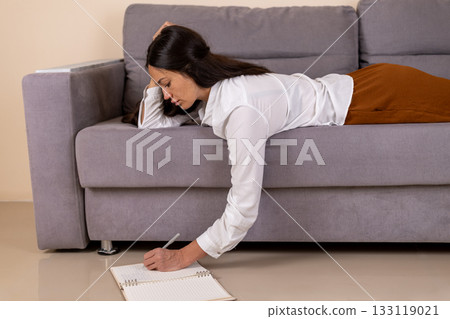 A woman is lying on the couch and writing in a notebook 133119021