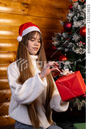 A young woman in a Santa hat sits by the Christmas tree, holding a red gift box 133119025
