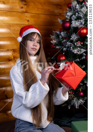 Girl in Santa hat opens present box by Christmas tree Girl in Santa hat opens present box by Christmas tree 133119033