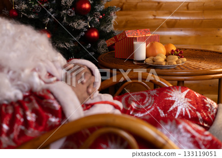 Santa Claus is sleeping in a chair next to the Christmas tree Santa Claus is sleeping in a chair next to the Christmas tree 133119051