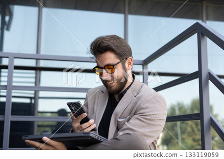 A man with a beard and sunglasses is sitting outdoors, looking at his smartphone A man with a beard and sunglasses is sitting outdoors, looking at his smartphone 133119059