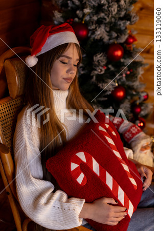 woman is sitting by a Christmas tree, holding a festive red pillow. 133119060