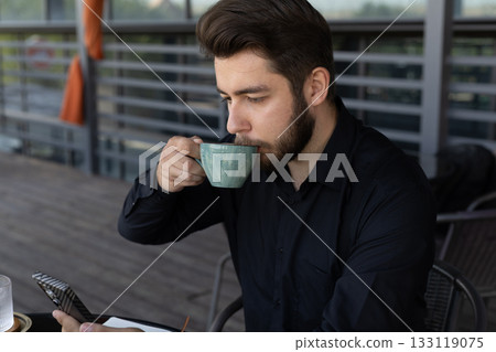 A bearded man is sitting and drinking from a cup while looking at his phone. 133119075