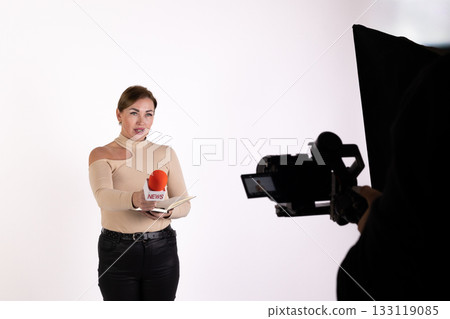 A reporter is standing in a studio holding a microphone as a camera records her 133119085