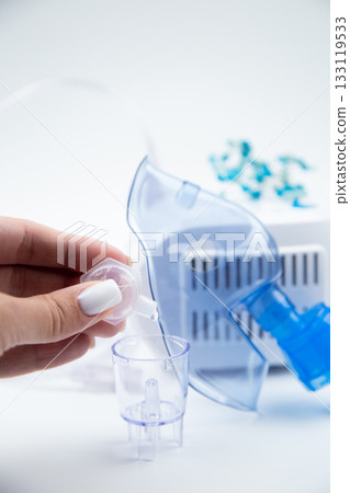 Vertical close-up photo of a medicinal solution dripping from an ampoule into an inhalation bowl against the background of a blue breathing mask and a device on a white background. The concept of 133119533