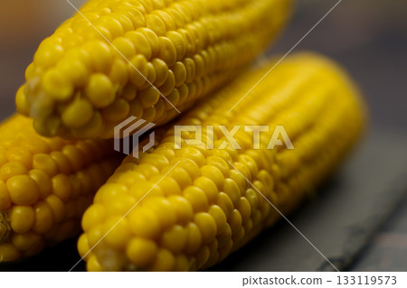 Close-up of three yellow peeled ears of corn with large grains lying on a black stone plank on a dark background with a focus in the center. The concept of healthy nutrition, natural iron, corn Close-up of three yellow peeled ears of corn with large grains lying on a black stone plank on a dark background with a focus in the center. The concept of healthy nutrition, natural iron, corn 133119573