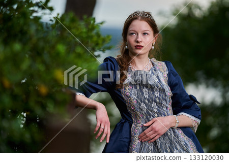 Portrait of a young redhead girl in Victorian style dress posing beside castle 133120030