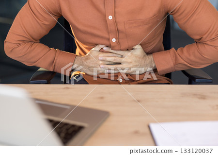 Young professional clutching his abdomen in pain at an office desk, struggling with indigestion or stomachache while trying to work, stressed and feeling unwell 133120037