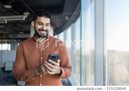 Indian man smiling and interacting with his mobile phone, standing in a modern office space with large windows, representing business communication and digital technology Indian man smiling and interacting with his mobile phone, standing in a modern office space with large windows, representing business communication and digital technology 133120048