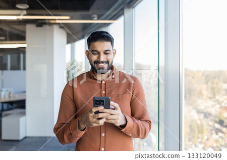 Indian businessman smiling while checking messages on his smartphone, connecting with people, and managing business in a contemporary office building with large windows Indian businessman smiling while checking messages on his smartphone, connecting with people, and managing business in a contemporary office building with large windows 133120049