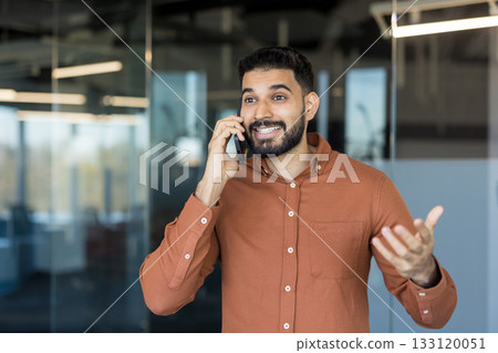 Young businessman smiling and talking on his mobile phone while gesturing in a modern office, conveying confident, cheerful communication, networking and professional connection 133120051