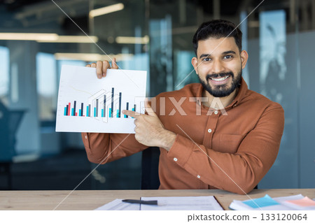 Smiling young indian businessman holding up a graph and pointing, explaining financial statistics during a presentation in a modern office setup, analyzing market trends Smiling young indian businessman holding up a graph and pointing, explaining financial statistics during a presentation in a modern office setup, analyzing market trends 133120064