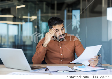 Young businessman at his office desk reading a troubling document, visibly frustrated and anxious over financial problems, paperwork and a looming business crisis Young businessman at his office desk reading a troubling document, visibly frustrated and anxious over financial problems, paperwork and a looming business crisis 133120066