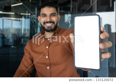 Smiling young adult man holding a modern smartphone with a clean white display for mockup design, presenting new technology in a professional office environment 133120072