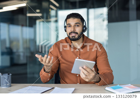 Young man wearing a headset with a microphone sitting at a desk, looking at the camera and gesturing while holding a digital tablet, representing online support and communication 133120079