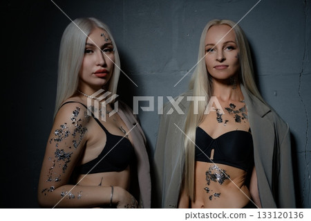 Two models in silver body art pose in stylish outfits on a dark backdrop Two models in silver body art pose in stylish outfits on a dark backdrop 133120136