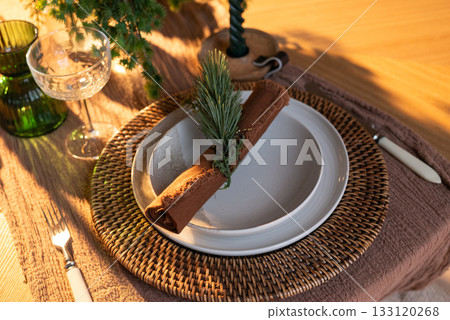 Festive table setting with woven charger, ceramic plates, rustic napkin and greenery decoration in warm holiday lighting. 133120268