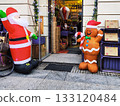 Inflatable Santa Claus and gingerbread man decorations stand outside a shop entrance. Santa, in his traditional red outfit, waves with one hand 133120484