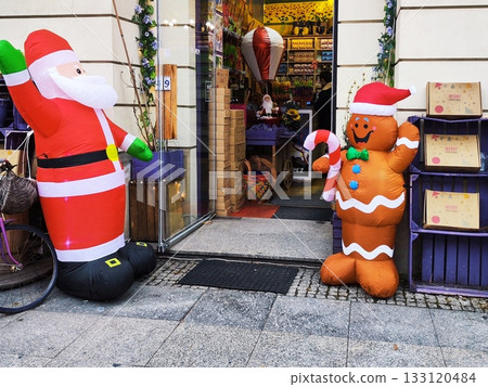 Inflatable Santa Claus and gingerbread man decorations stand outside a shop entrance. Santa, in his traditional red outfit, waves with one hand Inflatable Santa Claus and gingerbread man decorations stand outside a shop entrance. Santa, in his traditional red outfit, waves with one hand 133120484