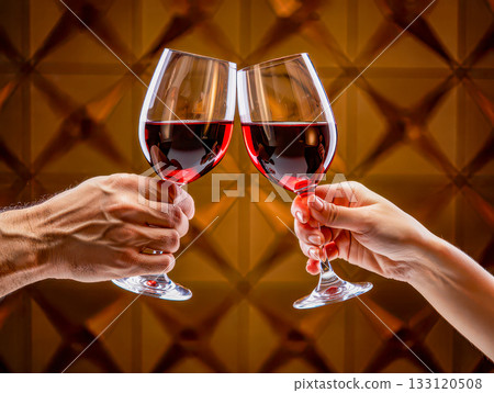 Romantic toast with red wine against a golden background. Two hands holding wine glasses clink together, filled with rich red wine.  133120508