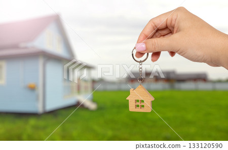 Key to new home: woman's hand holding wooden house keychain with blurred background Key to new home: woman's hand holding wooden house keychain with blurred background 133120590