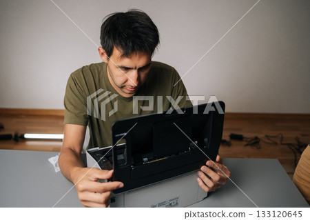Top view of technician examining components of disassembled laser printer, troubleshooting and performing maintenance in workshop environment, sitting at table on isolated background. 133120645
