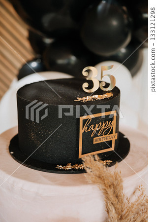 Black birthday cake with gold 35 topper on pedestal with pampas decor at elegant party 133121298