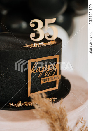 Black birthday cake with gold 35 topper and plaque, pampas decor and balloons background 133121300