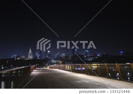 A nighttime view of Warsaw skyline captured from a modern footbridge, with shining skyscrapers and the Palace of Culture glowing in the distance. 133121448