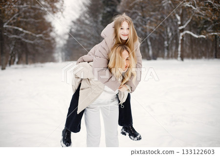 Mother and daughter having fun in forest on snow day. Family wearing beige warm jackets. Woman carrying girl piggyback. 133122603