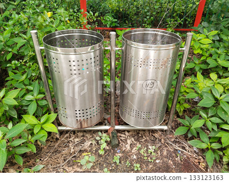 Stainless steel double dustbin unit in outdoor park, cylindrical perforated design for public sanitation, surrounded by green foliage, promotes cleanliness and responsible waste disposal in public Stainless steel double dustbin unit in outdoor park, cylindrical perforated design for public sanitation, surrounded by green foliage, promotes cleanliness and responsible waste disposal in public 133123163