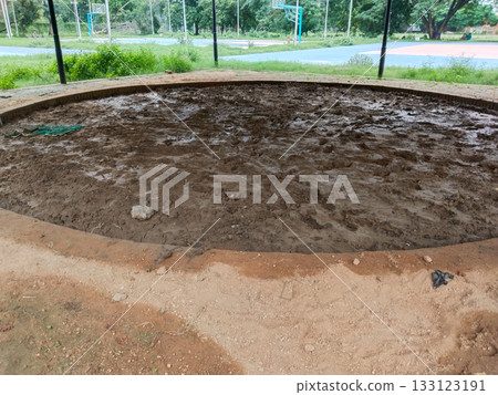 Muddy circular sports arena with wet soil surface and brick boundary in open park, traditional outdoor ground prepared for local games or wrestling, natural earth field surrounded by green landscape 133123191