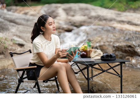 Reading in Nature. Young woman enjoying a book by a tranquil stream, promoting relaxation and escape. Reading in Nature. Young woman enjoying a book by a tranquil stream, promoting relaxation and escape. 133126061