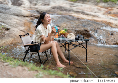 Joyful Moments. Young woman laughing with a cup of tea by a flowing stream, embracing happiness and nature. 133126064