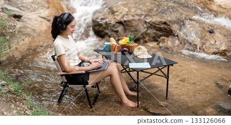 Digital Nomad. Young woman working on a laptop by a stream, combining productivity and nature. Digital Nomad. Young woman working on a laptop by a stream, combining productivity and nature. 133126066