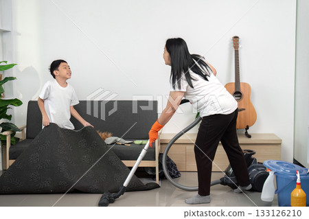 A young boy plays while his parent vacuums the living room, highlighting family engagement in cleaning tasks.. 133126210