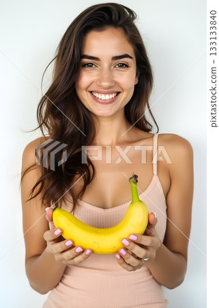 Smiling woman holding a ripe banana in a bright indoor setting with a neutral background 133133840