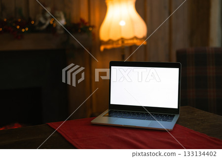 Laptop with white screen mockup on a table with a red runner in a cozy, dimly lit interior with a fireplace and vintage lamp 133134402