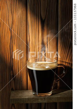 Ray of light falls on transparent cup of coffee standing on shelf against wooden background Ray of light falls on transparent cup of coffee standing on shelf against wooden background 133137968