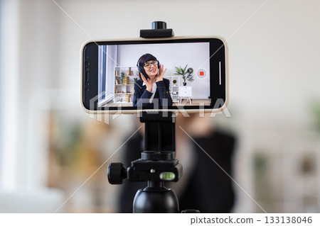 Caucasian woman sitting at table filming a video blog about relaxation techniques, wearing headphones, demonstrating calming exercises, modern workspace, professional setup. Caucasian woman sitting at table filming a video blog about relaxation techniques, wearing headphones, demonstrating calming exercises, modern workspace, professional setup. 133138046