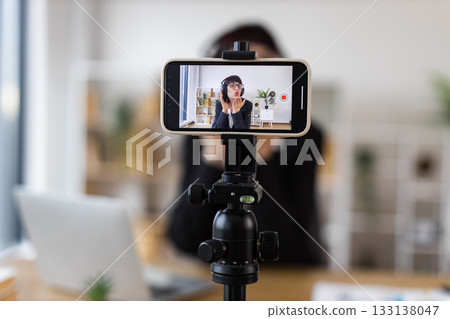 Caucasian woman sitting at table filming a video blog about relaxation techniques, wearing headphones, demonstrating calming exercises, modern workspace, professional setup. Caucasian woman sitting at table filming a video blog about relaxation techniques, wearing headphones, demonstrating calming exercises, modern workspace, professional setup. 133138047