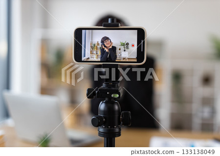Young woman in modern office environment recording video with smartphone on tripod, appearing relaxed and engaging. Young woman in modern office environment recording video with smartphone on tripod, appearing relaxed and engaging. 133138049