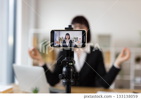Caucasian woman in office environment recording meditation video at desk, combining work and relaxation. Professional appearance demonstrates how to balance workplace stress using mindfulness. 133138059
