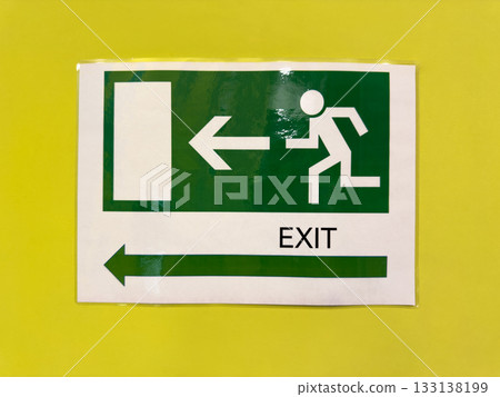 Green sign on yellow wall with building exit direction. Safe navigation indoors in case of fire or accident. Street exit sign with directional arrow and symbol of person running out Green sign on yellow wall with building exit direction. Safe navigation indoors in case of fire or accident. Street exit sign with directional arrow and symbol of person running out 133138199