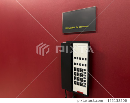 Landline telephone on red wall with warning sign directing you to operator. Emergency communication within building in case of problems or accidents. Phone with communication buttons Landline telephone on red wall with warning sign directing you to operator. Emergency communication within building in case of problems or accidents. Phone with communication buttons 133138206