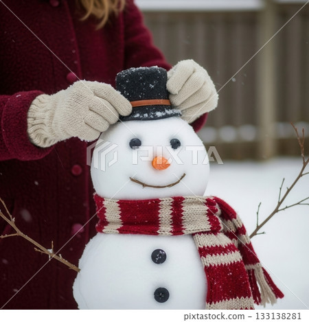 Child adjusts the hat of a snowman wearing a red scarf during a snowy winter day, capturing the warmth and joy of holiday moments, ideal for festive lifestyle photography and seasonal campaigns. Child adjusts the hat of a snowman wearing a red scarf during a snowy winter day, capturing the warmth and joy of holiday moments, ideal for festive lifestyle photography and seasonal campaigns. 133138281