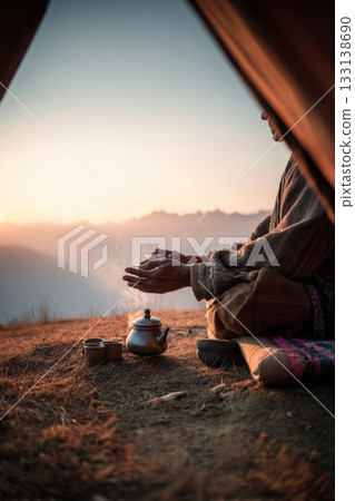 Man warming hands over steaming teapot at sunrise near tent, peaceful morning in the mountains ideal for travel, mindfulness, camping solitude, and slow outdoor living imagery. 133138690